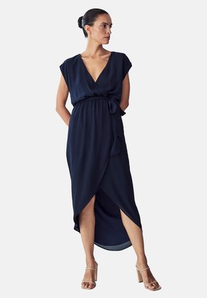 Robe portefeuille bleu marine avec un décolleté en V profond, des manches courtes, et une taille nouée, présentant une fente latérale et un tissu fluide. Portée avec des talons beiges.