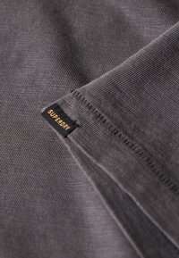 Tela gris con una textura suave, que presenta una etiqueta cosida que dice "SUPERDRY" en negro y dorado. El diseño es minimalista y limpio.