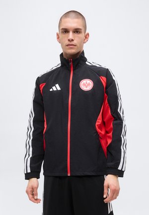 Jeune homme aux cheveux courts portant une veste de sport noire et rouge avec des bandes blanches et un logo d'aigle sur la poitrine, debout devant un fond uni.