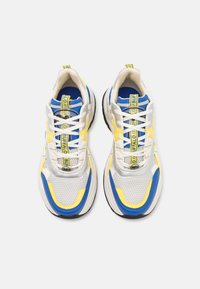 Sneaker atletiche bianche e blu con parte superiore in rete, accenti gialli, dettagli argentati e una suola nera spessa con motivi testurizzati.