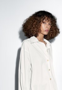 Světlé šedé košile na knoflíky s límečkem, zdobené zlatými knoflíky a hladkou texturou, nošené modelem s kudrnatými vlasy.