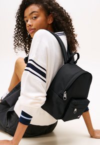 Mochila de nylon negro con bolsillos con cremallera, correas acolchadas y forma redondeada. Llevada por una persona con un suéter blanco con rayas marinas.
