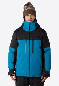 Blå och svart vattentät jacka med hög krage, justerbar huva, framdragkedjor och elastiska ärmslut. Kombinerad med en brun stickad mössa.