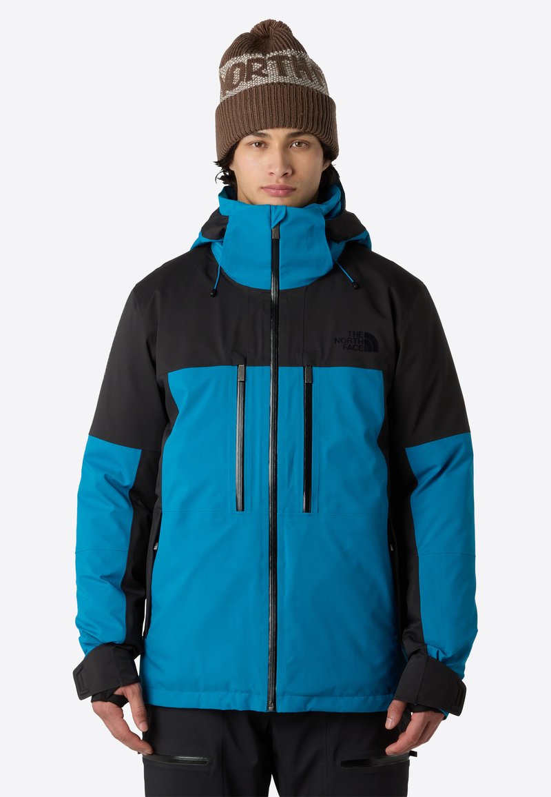 Blå och svart vattentät jacka med hög krage, justerbar huva, framdragkedjor och elastiska ärmslut. Kombinerad med en brun stickad mössa.