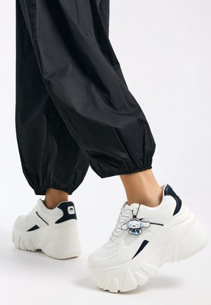 Weiße Plateau-Sneaker mit strukturierter Oberfläche, blauen Akzenten und einem Charms-Anhänger. Kombiniert mit schwarzen, schmal zulaufenden Hosen.