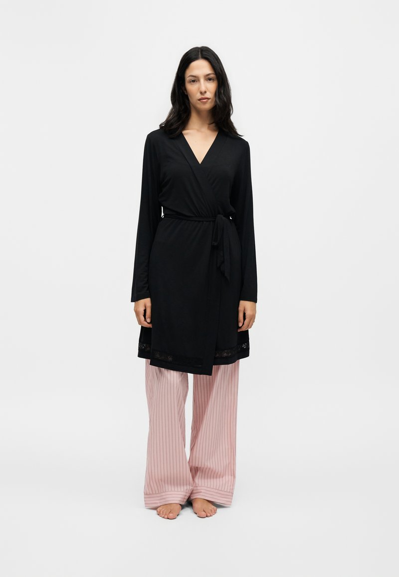 Γυναίκα με μακριά σκούρα μαλλιά, φοράει μαύρο φόρεμα τύπου wrap πάνω από ροζ ριγέ φαρδιά παντελόνια, στέκεται ξυπόλητη μπροστά σε απλό φόντο.