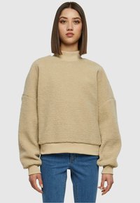 Sweatshirt en polaire beige avec un col haut, des épaules tombantes et des manches larges. Surface texturée avec une coupe légèrement cropped. Associé à un jean bleu.