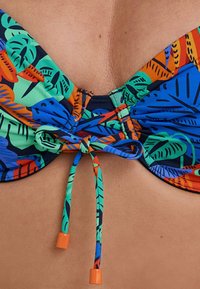 Top bikini con vivaci motivi a foglia nei colori verde, arancione e blu. Presenta una chiusura centrale con laccetti arancioni e dettagli in tessuto arricciato.