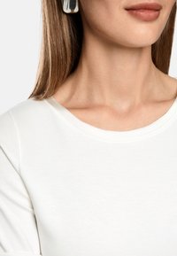 White cotton top with a round neckline, visible stitching, and short sleeves. Silver statement earrings add contrast to the light fabric.