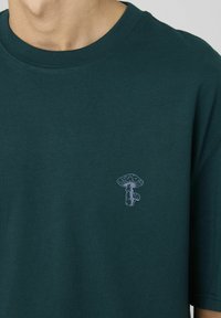 T-shirt en coton vert foncé avec un col rond et des manches courtes, arborant un graphique de champignon blanc sur la poitrine gauche.