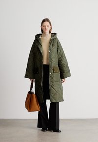 Khaki zelený prošívaný dlouhý kabát s kapucí, který má dvě přední kapsy a zlaté detaily. Nošený s béžovým svetrem a černými kalhotami.