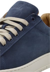 Navy blue suede sneaker featuring beige laces, round eyelets, and a contrasting light-colored rubber sole. Textured upper with embossed logo.