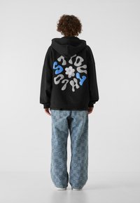 Sudadera negra con diseño de texto abstracto colorido en la parte de atrás, combinada con pantalones vaqueros holgados de color azul claro con patrón. Zapatillas blancas visibles.