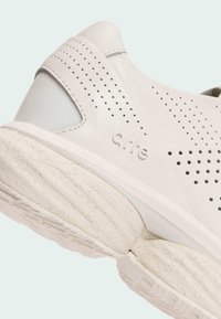 Baskets sportives blanches avec cuir perforé, accents lisses et semelle texturée. Marque visible sur le côté, mettant en avant un design minimaliste.