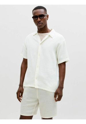 Homme noir portant des lunettes de soleil rectangulaires et une chemise blanche texturée à manches courtes avec des boutons, assortie à un short, sur un fond uni.