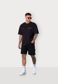 Black oversized t-shirt with white text, paired with black cargo shorts. White sneakers and sunglasses complete the look. Smooth cotton fabric.