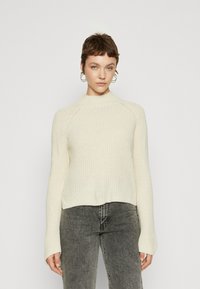 Maglione in crema lavorato a maglia con colletto alto, texture a costine e maniche lunghe, abbinato a jeans grigi scuri. Design semplice e moderno.