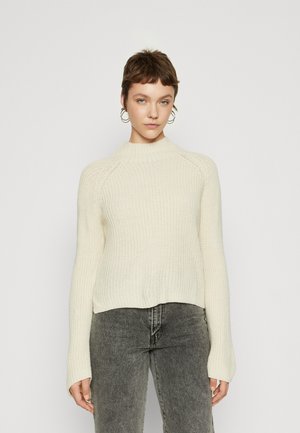 Pull en tricot crème avec un col montant, texture côtelée et manches longues, associé à un jean gris foncé. Design simple et moderne.