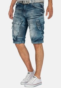 Uomo che indossa pantaloni shorts in denim blu scolorito con tasche cargo etichettate "CIPO" e "BAXX", sneakers bianche e una cintura grigia.