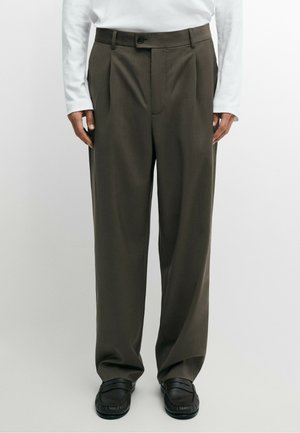 Person wearing dark brown wide-leg trousers, black loafers, and a white long-sleeve top, standing against a plain light background.