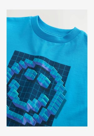 Világoskék pamut póló, sötét rács háttérrel, amely 3D pixel dizájnnal rendelkezik türkiz és lila színekben. Kerek bordázott nyakkivágás.