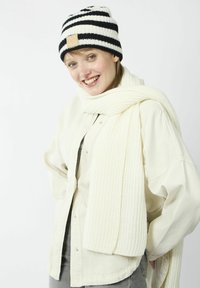 Gehaakte zwart-wit gestreepte beanie met een tan lederen patch; dikke witte geribde sjaal; lichtbeige overhemd met knopen; grijze broek.