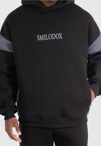 Schwarzer Hoodie mit grauen Akzenten und gesticktem "SMILODOX"-Logo auf der Brust. Weicher Stoff, lockere Passform, gerippte Bündchen und Saum.
