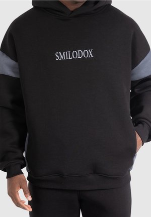 Černá mikina s šedými akcenty a vyšívaným logem "SMILODOX" na hrudi. Měkká tkanina, uvolněný střih, žebrované manžety a lem.