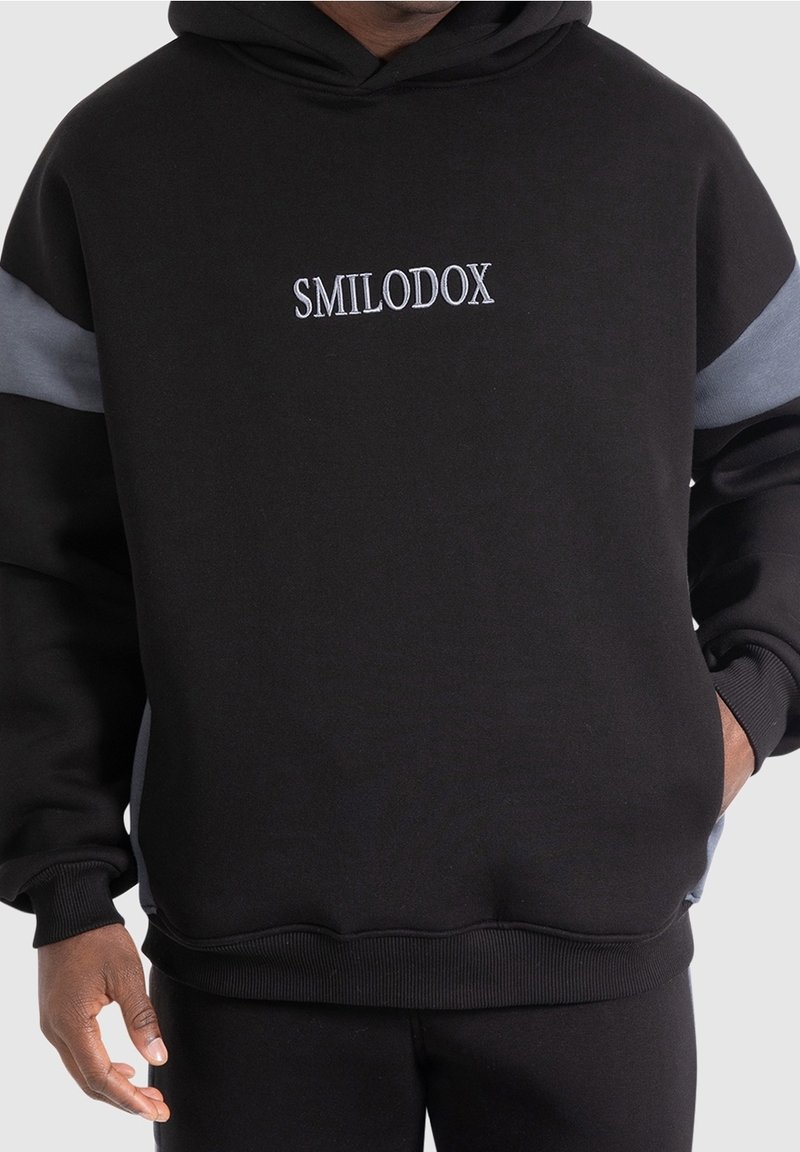 Schwarzer Hoodie mit grauen Akzenten und gesticktem "SMILODOX"-Logo auf der Brust. Weicher Stoff, lockere Passform, gerippte Bündchen und Saum.