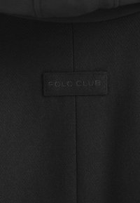 Svart tygmärke med "POLO CLUB" präglat i mörkgrått. Texturen verkar slät och något matt. Positionerat inom plaggets söm.