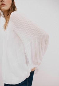 Hvid, plisseret bluse med lange, voluminøse ærmer. Blødt, letvægtsstof med en afslappet pasform, båret over denimbukser.