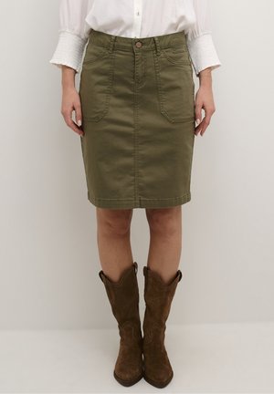 Femme portant une jupe mi-longue vert olive, un chemisier blanc avec des poignets texturés et des bottes de cowboy en daim marron, debout sur un fond uni.