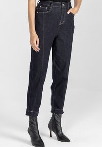 Granatowe dżinsy typu straight-leg z jasnym stębnowaniem, przednimi kieszeniami i podwiniętym dołem, noszone z czarnymi botkami na obcasie.