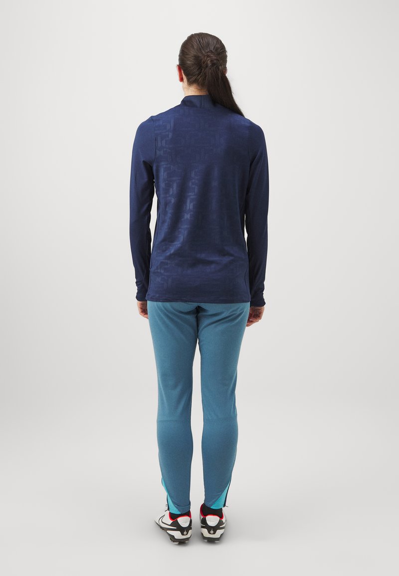 Marineblaues Langarmoberteil mit subtiler Struktur, kombiniert mit hellblauen, schmal zulaufenden Hosen und schwarz-weißen Sportschuhen.