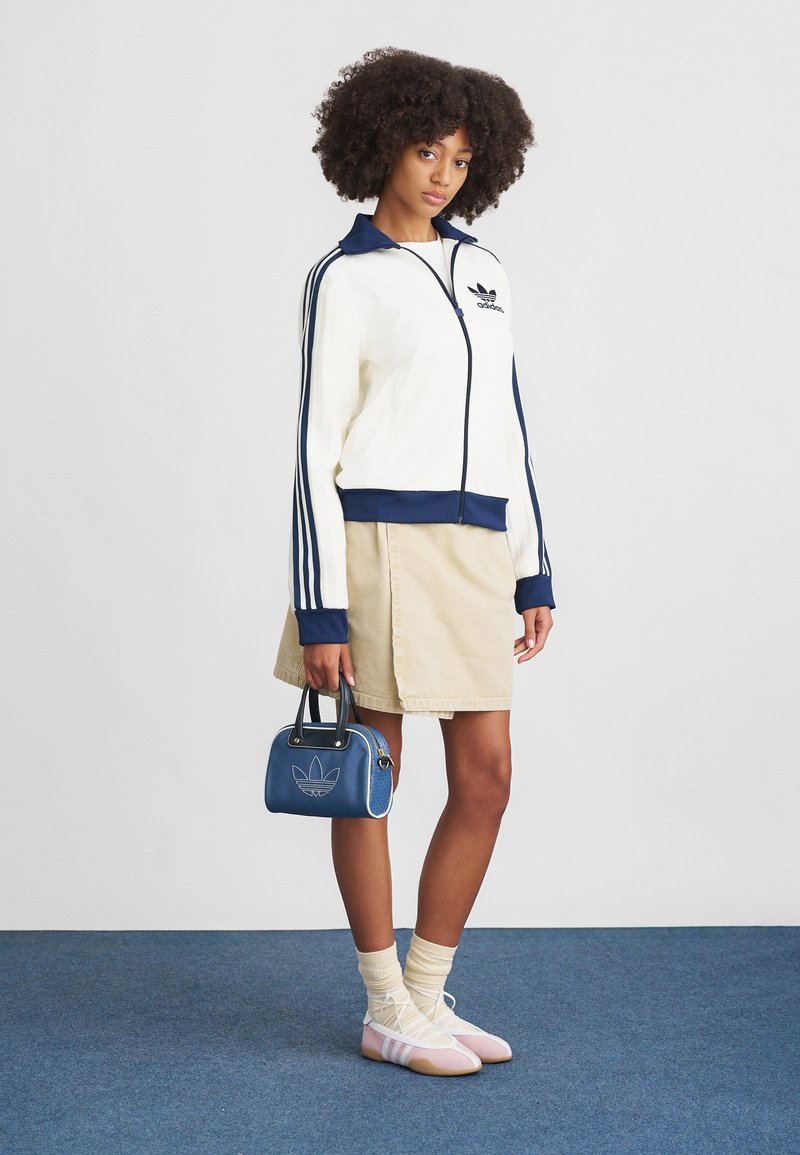 White zip-up jacket with navy accents and three stripes, beige skirt, blue handbag with logo, and pink footwear. Soft textures throughout.