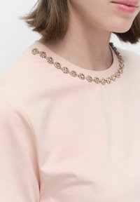 Ljusrosa topp med rundad halsringning prydd med en kedja av metalliska detaljer och små dekorativa inslag kring kragen.