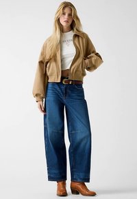 Bruine cropped jas over een wit shirt met logo, gecombineerd met wijde blauwe jeans en bruine enkellaarzen. Soepele texturen en een casual pasvorm.