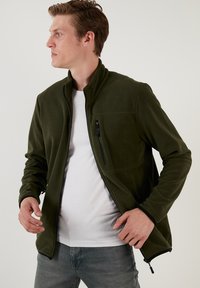 Giacca in fleece verde scuro con colletto alto, chiusura con zip frontale e tasca sul petto. Presenta una superficie testurizzata e cerniere nere a contrasto.