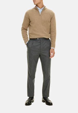 Beige Wollpullover mit Reißverschlusskragen, kombiniert mit grauen Hosen mit Knopf und Falten. Schwarze Slipper runden das Outfit ab.