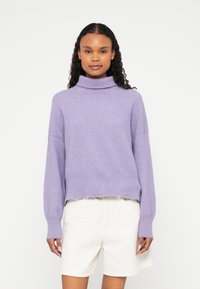 Lilakas augstu kakla džemperis no mīksta adīta auduma ar brīvu piegriezumu, nolaistām plecu līnijām un ribota auduma aprocēm un apakšmalu. Kombinēts ar gaišām šortiem.