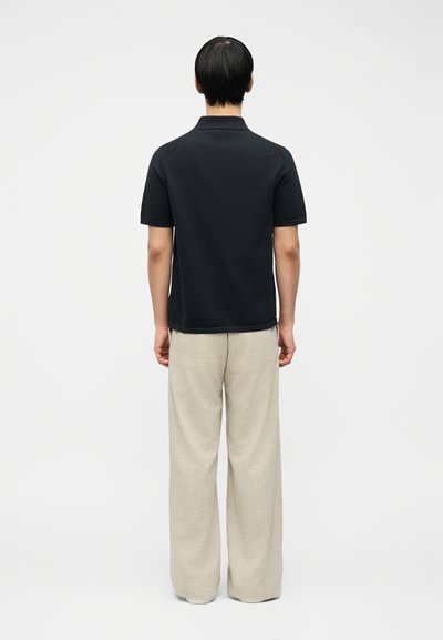 Άνδρας όρθιος με την πλάτη προς το φακό, φοράει μαύρο πόλο με κοντά μανίκια και μπεζ παντελόνι με φαρδιές πατούσες, με λευκό απλό φόντο.