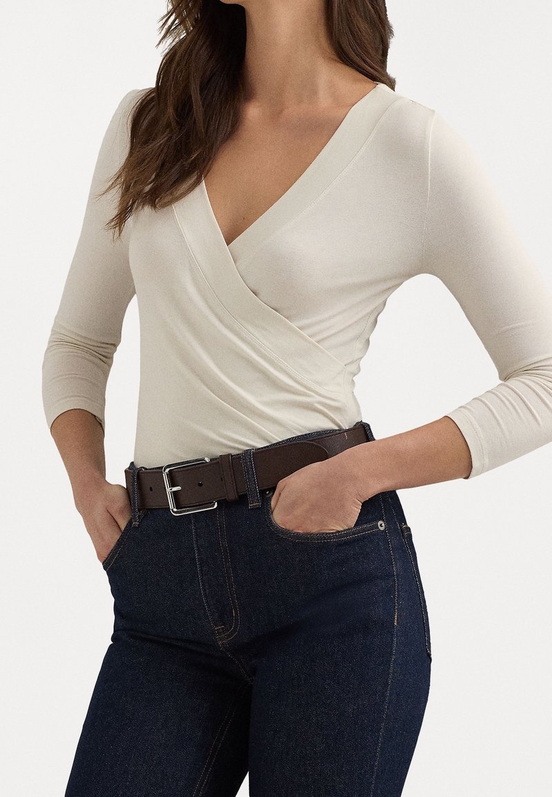 Long-sleeve wrap bodysuit in beige with a V-neck, paired with dark blue jeans and a brown belt featuring a metal buckle and textured leather.