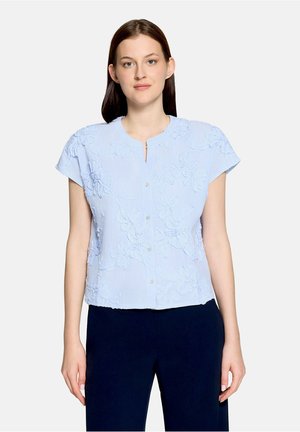 Femme portant un chemisier bleu clair à manches courtes avec broderie florale et un pantalon bleu marine foncé, debout sur un fond blanc.