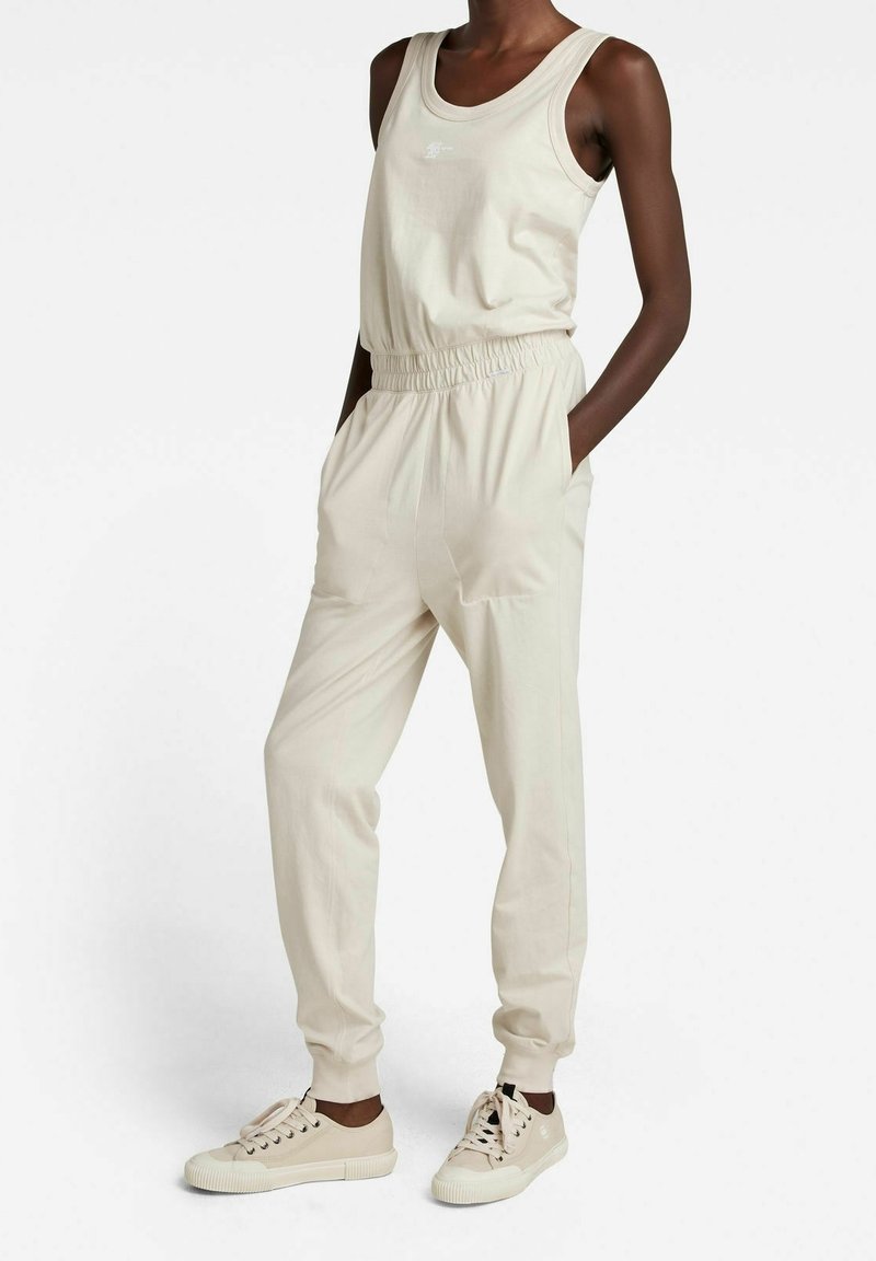 Smal person i beige linne, matchande joggingsbyxor med elastisk midja och beige snörskor, stående med händerna i fickorna.