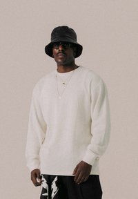 Suéter blanco con relieve y mangas largas, combinado con pantalones negros de estampado. Los accesorios incluyen un sombrero tipo bucket negro y collares de oro en capas.