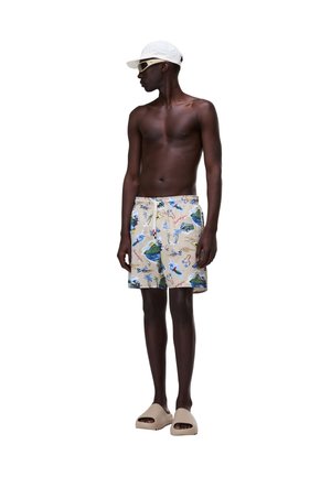 Uomo senza maglietta con pantaloncini da bagno beige stampati, cappellino bianco, sandali beige e occhiali da sole bianchi, in piedi e guarda a sinistra.