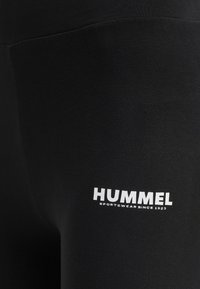 Leggings noirs en tissu extensible avec une texture lisse. Présente un logo blanc avec les mots "HUMMEL" et "SPORTSWEAR SINCE 1923."