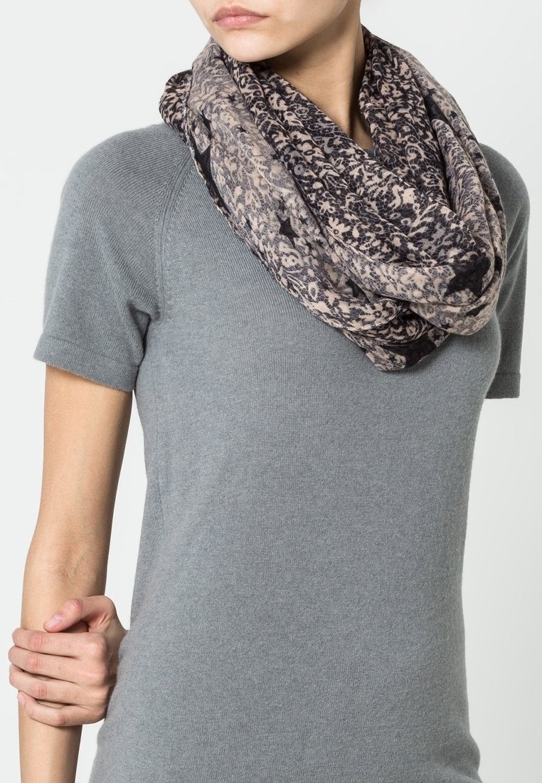 Femme portant une écharpe infinie beige et noire à motifs sur un haut en maille gris à manches courtes, une main tenant doucement son bras opposé.