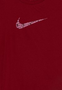 Crvena Nike Pro majica s prilagođenim dizajnom. Sadrži bijeli Nike swoosh logo i ponavljajući tekst "Nike Pro" u ružičastoj boji. Glatka tekstura.
