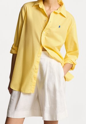 Persoon draagt een loszittend geel overhemd met opgerolde mouwen en witte shorts met hoge taille, één hand in de zak, tegen een effen achtergrond.